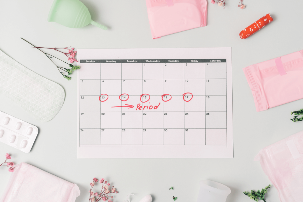 A calendar with multiple days circled indicating a woman's period. Sanitary pads and women's menstruation products are surrounding this calendar.