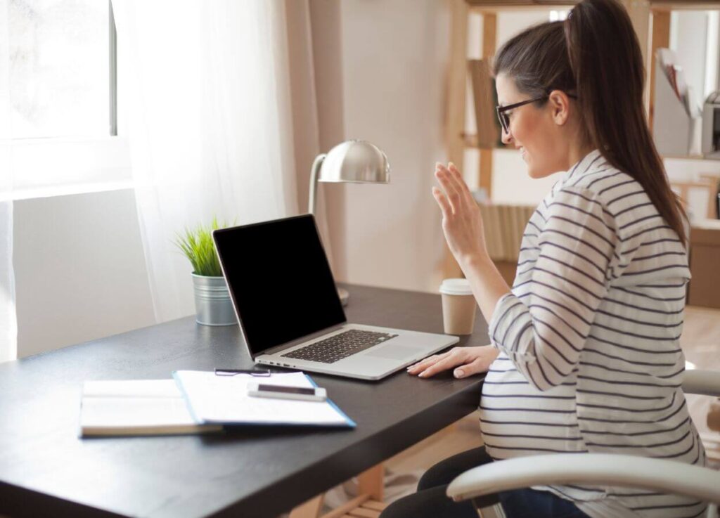 Pregnant college student sitting at a desk on a video call, smiling and waving to the camera