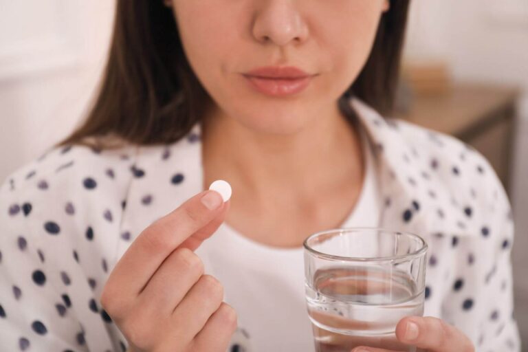 Woman holding a glass of water in one hand, and a small white pill in the other hand