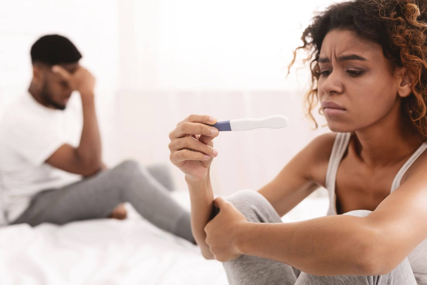 An upset woman looks at a pregnancy test, as her partner holds his head in the background