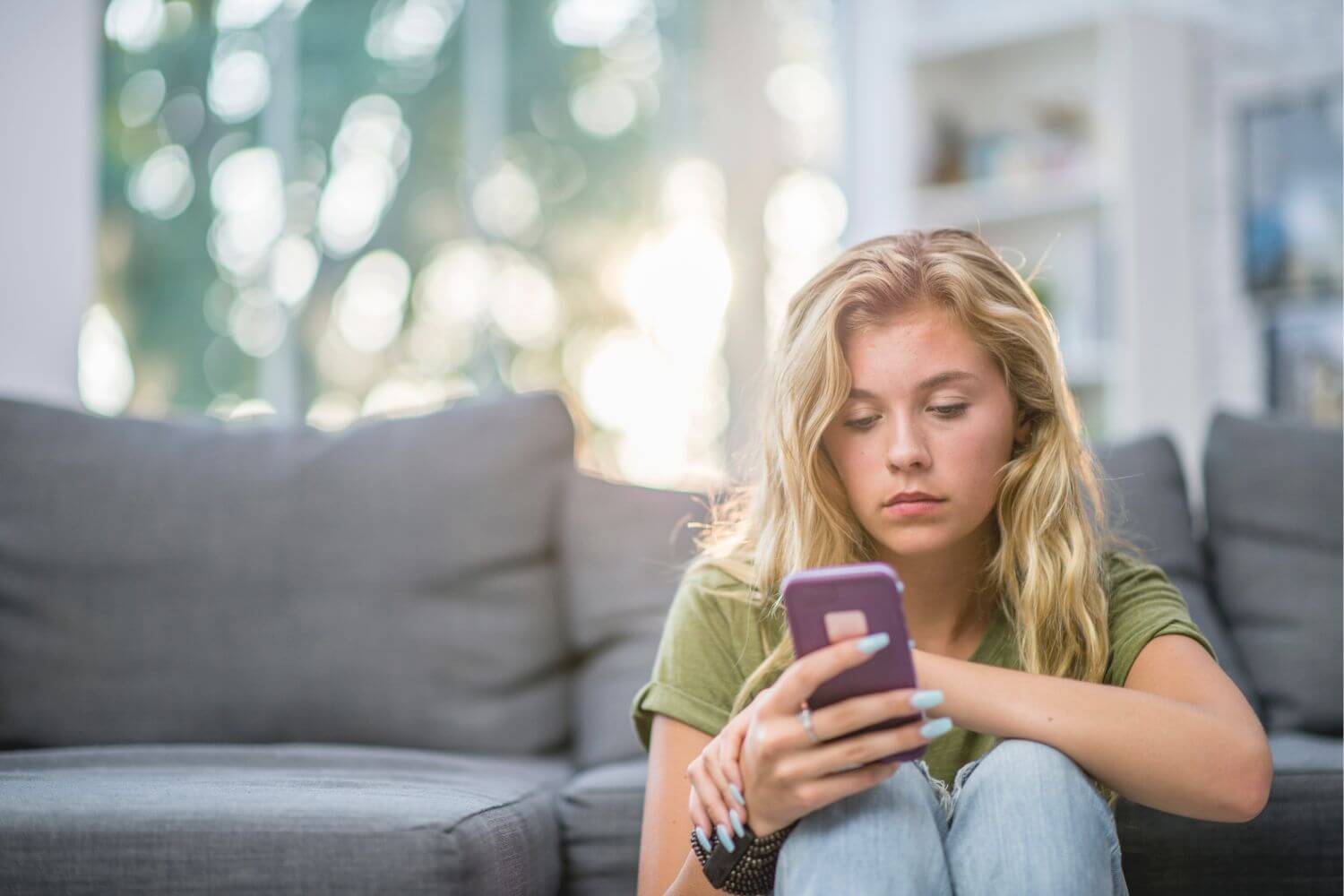 Woman sitting next to a couch, scrolling on her cell phone