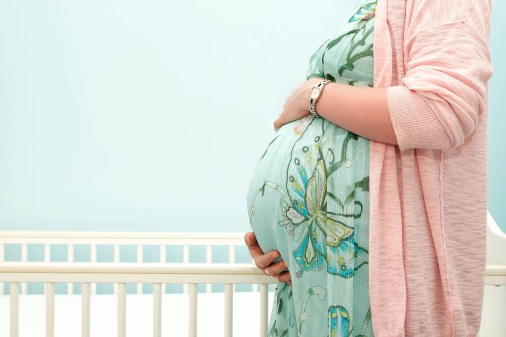 Pregnant woman in a floral dress and pink sweater standing next to a crib, holding her belly