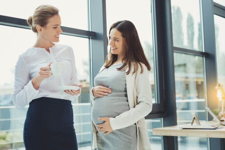 Pregnant employee holding her belly as she speaks with an associate at the workplace
