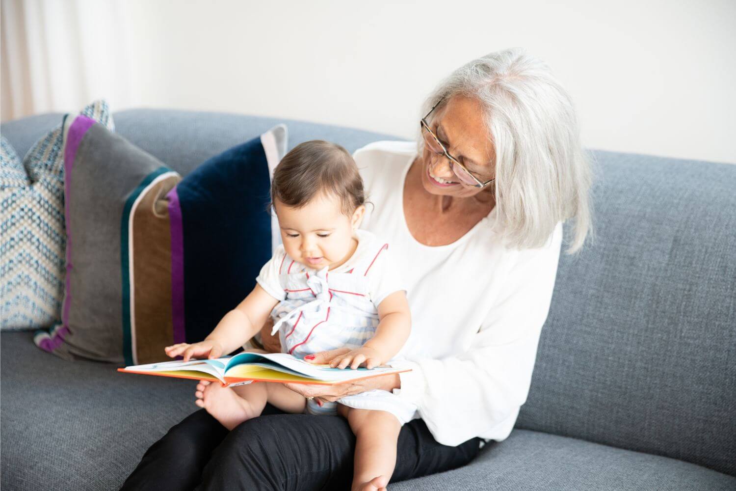 Grandma is holding a baby on her lap while sitting on a grey couch