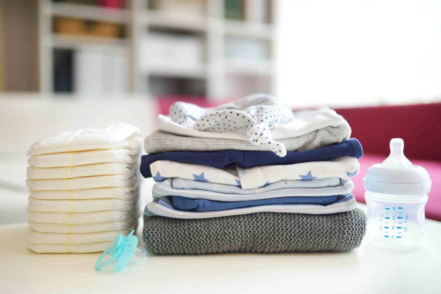 A stack of baby diapers, baby clothes, and a bottle are placed on a table in a home