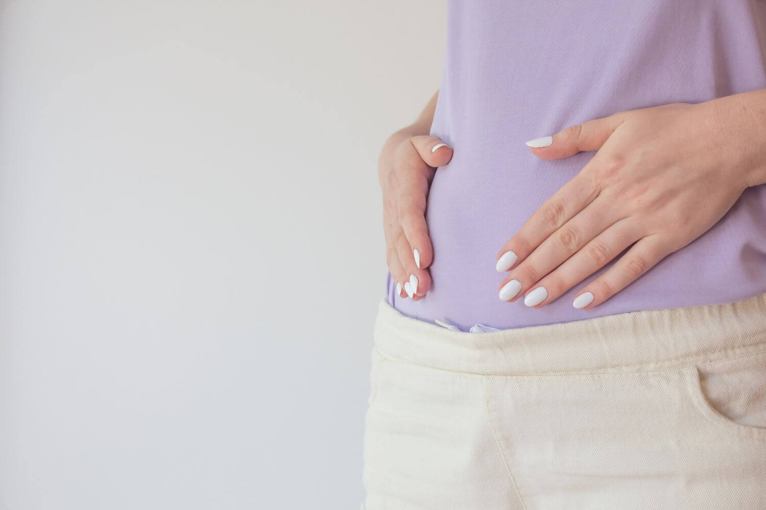 woman wearing a purple shirt and white pants holds her hands over her stomach