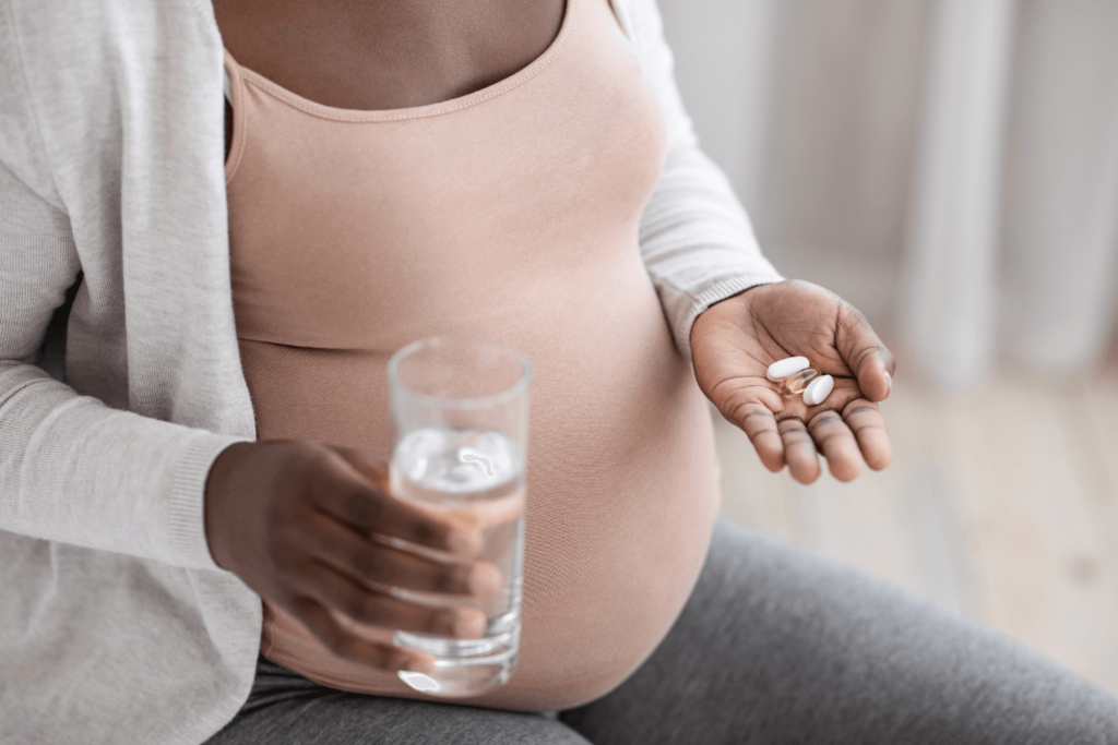 Pregnant woman holding a glass of water in one hand, and a variety of vitamins in the other