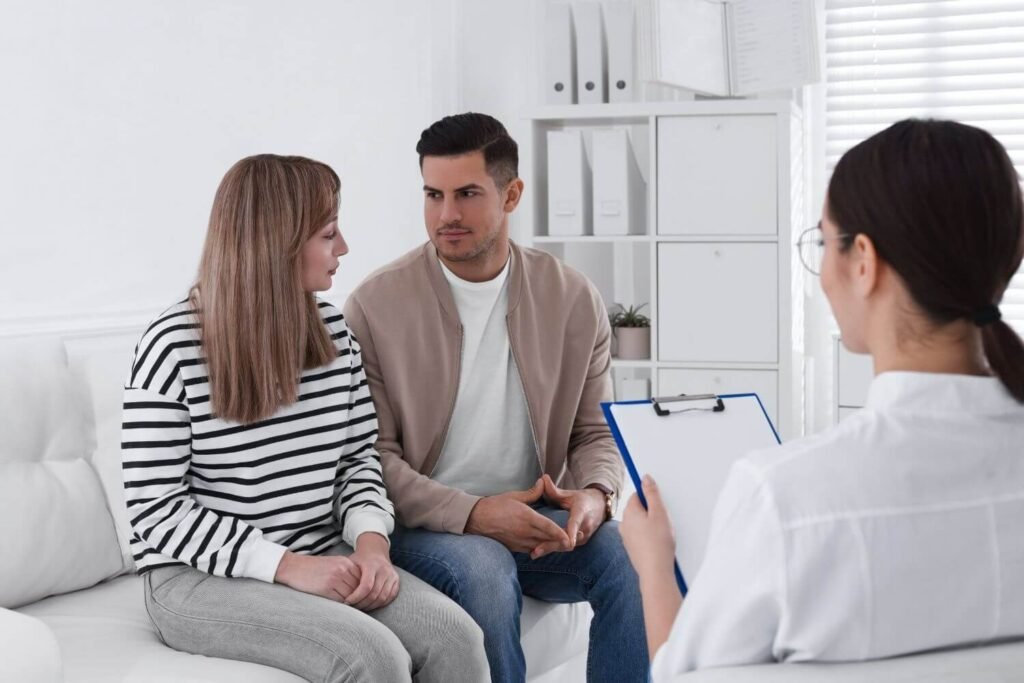 A young couple is sitting on a couch across from a physician holding a clip board